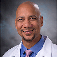 Headshot of a smiling man in a suit, tie and white Doctor's jacket against a gray background.