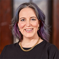 Headshot of a smiling woman in a black top and gold necklace, against a wood background with a window.