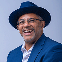 Headshot of a smiling man in a blue suit and white shirt, with a hat and eye glasses, against a blue background.