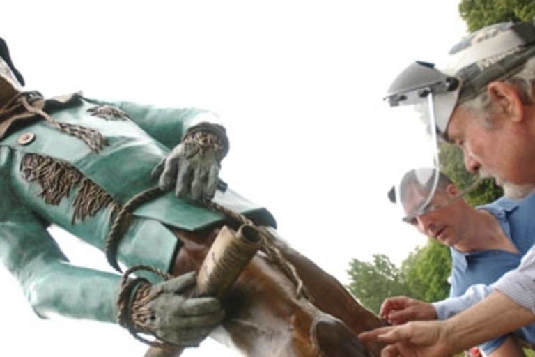 Large bronze statue of a cowboy being examined by two men.
