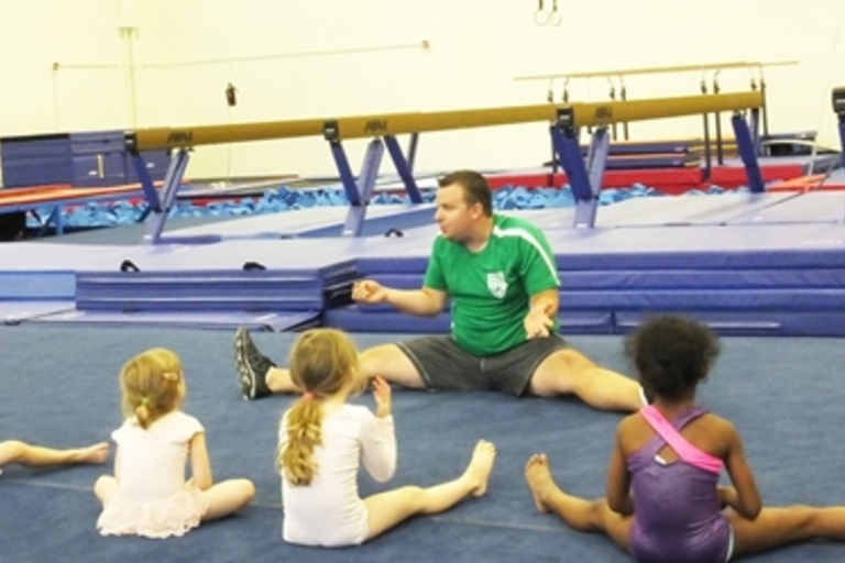 Gymnastics coach leads four young children in stretching exercises.
