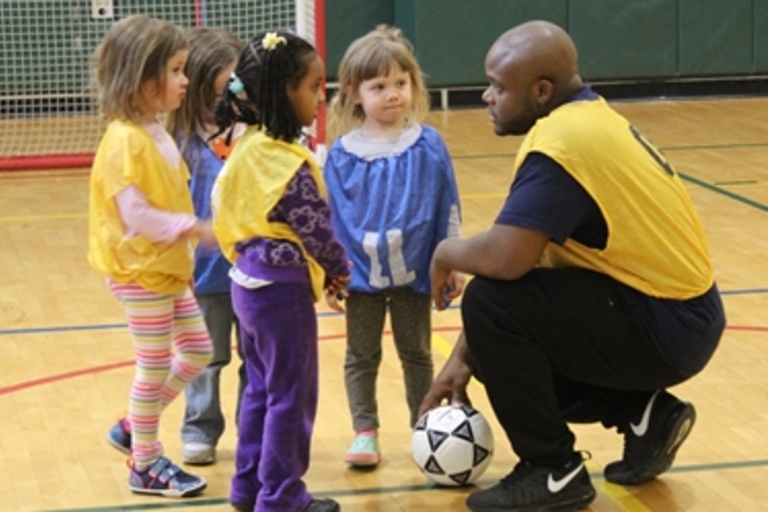 Coach kneels with soccer ball, talking to preschoolers in gym.
