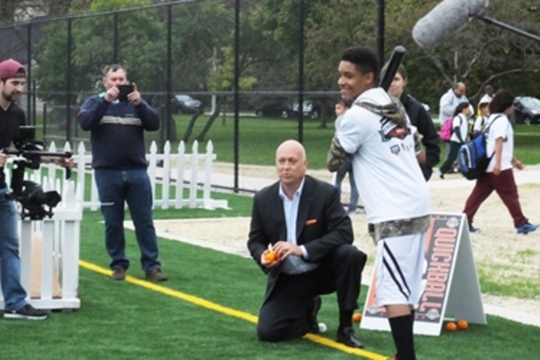 Teenager about to hit a quickball pitched by a man, as two others film the scene.