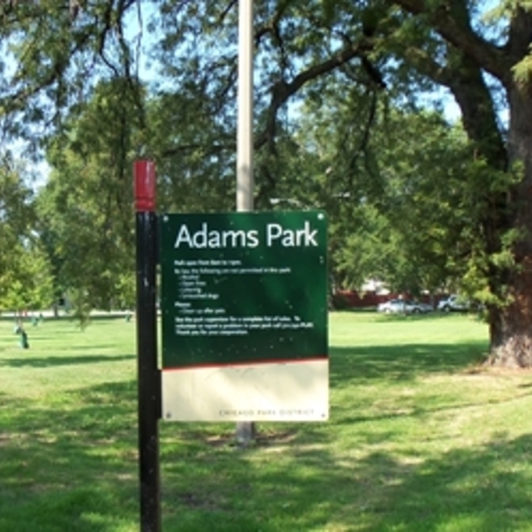 Adams Park sign in a grassy park with trees and sidewalks.