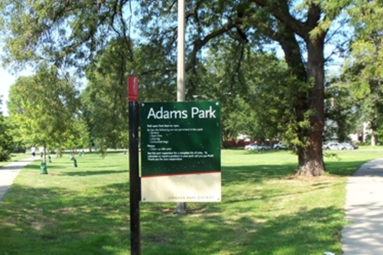 Adams Park sign in a grassy park with trees and sidewalks.