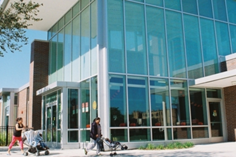 Modern library building with glass facade. Parents with strollers walk outside.