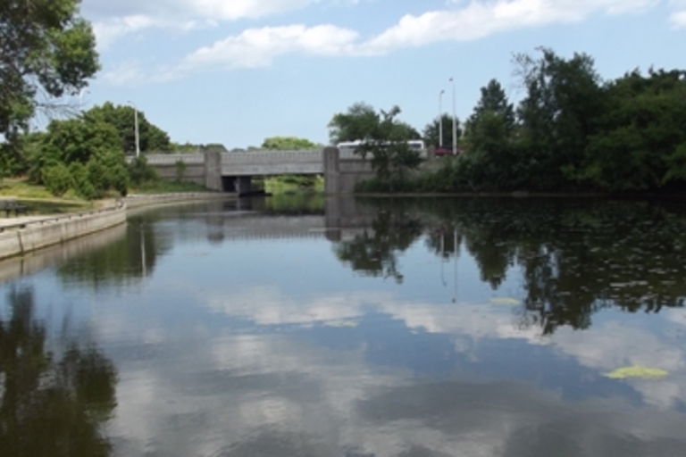 A calm river with a bridge in the background. Trees and a paved walkway line the riverbank.