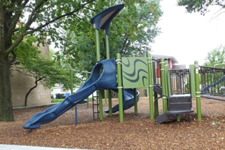 Playground with blue slide and green climbing structure. Wood chip ground cover.