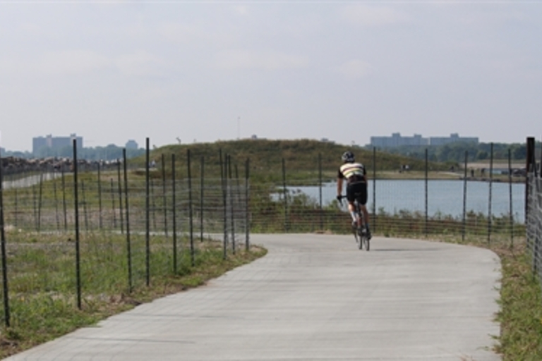 Cyclist rides away from camera on paved path next to water.