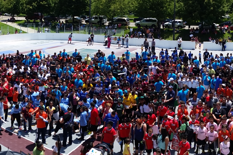 Large group of children and adults wearing brightly colored shirts gathered on an outdoor court.