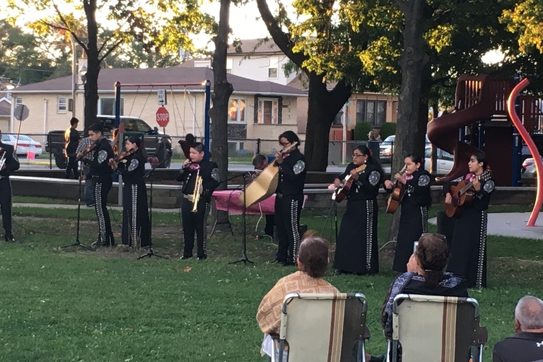 Mariachi band plays in a park. People sit in lawn chairs watching them.