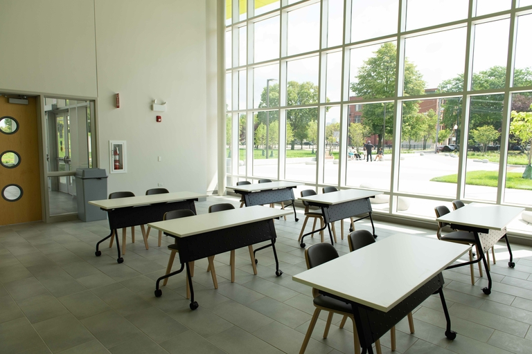Classroom with white tables and dark chairs by large windows showing a park.