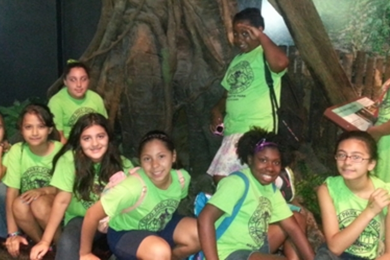Group of girls in matching green shirts pose by a large tree root.
