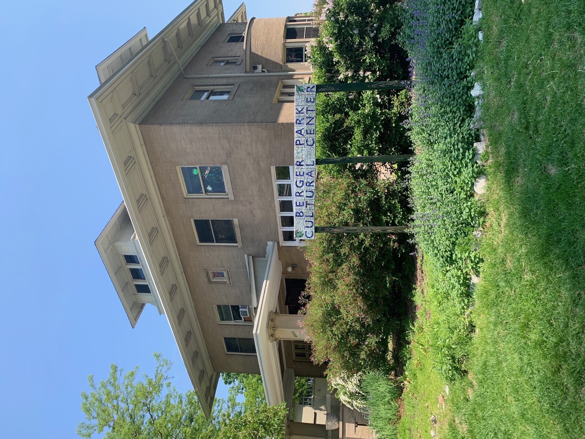 Berger Park Cultural Center, a tan brick building with white trim, stands angled against a clear blue sky. Lush greenery and a mosaic sign mark the entrance.