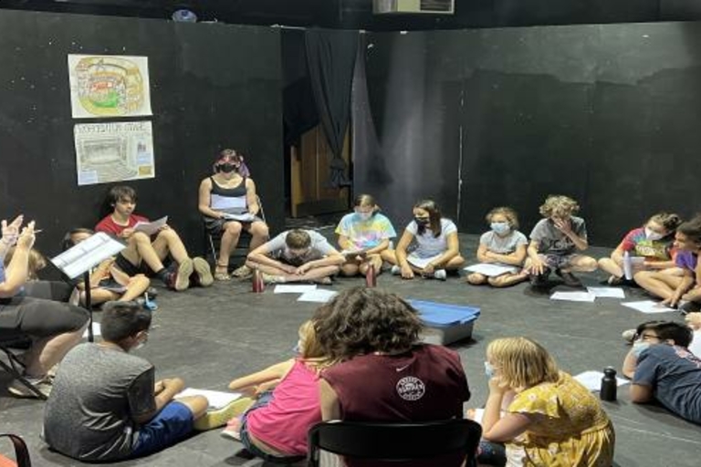 Children sit in a circle on a black floor in a theatre, reading scripts with an instructor.  Drawings of theatre stages are tacked to the back wall.