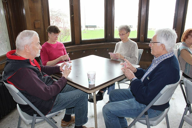 Four seniors play cards at a square table in a room with large windows.