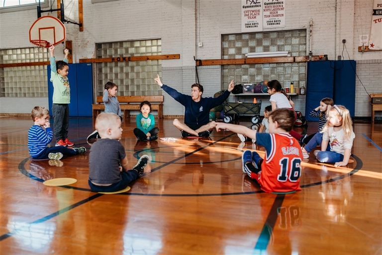 Children sit on a gym floor while a coach gives instructions. One child raises their hand.