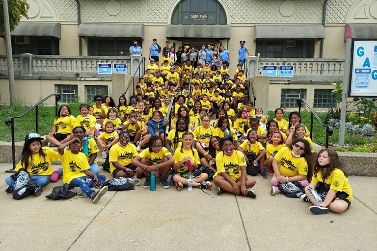 Large group of children and adults in matching yellow shirts pose outside Gage Park field house.
