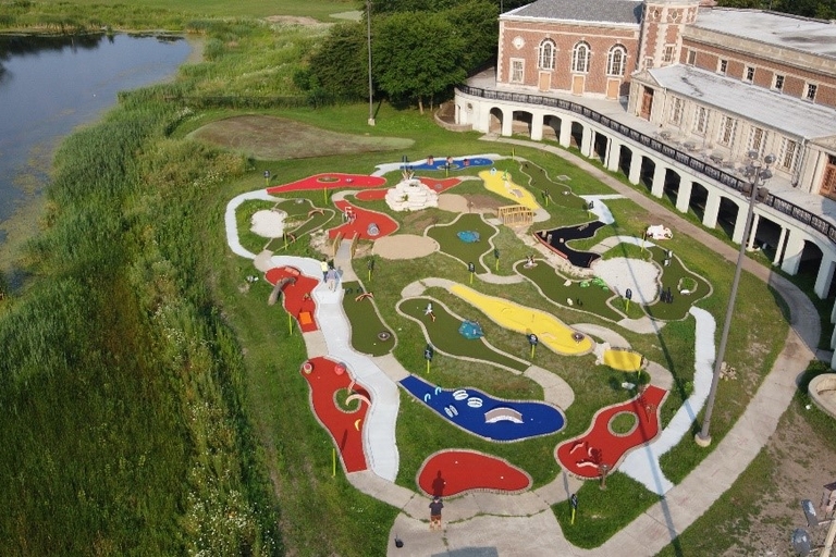 Aerial view of a colorful mini golf course next to a historic building and a pond.
