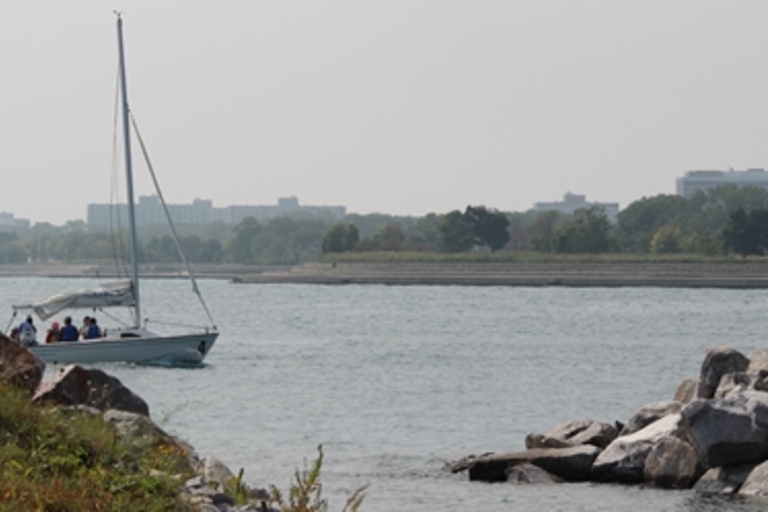 Sailboat with people on calm water near rocky shore. Cityscape in distance.
