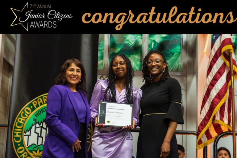 Three women at an awards ceremony, one holding an award, in front of a Chicago Parks banner and US flag.