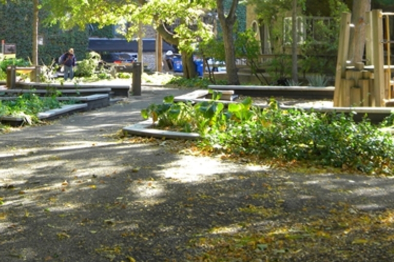Community garden with paved paths, raised planting beds, and a wooden play structure. Fallen yellow leaves scatter the ground.