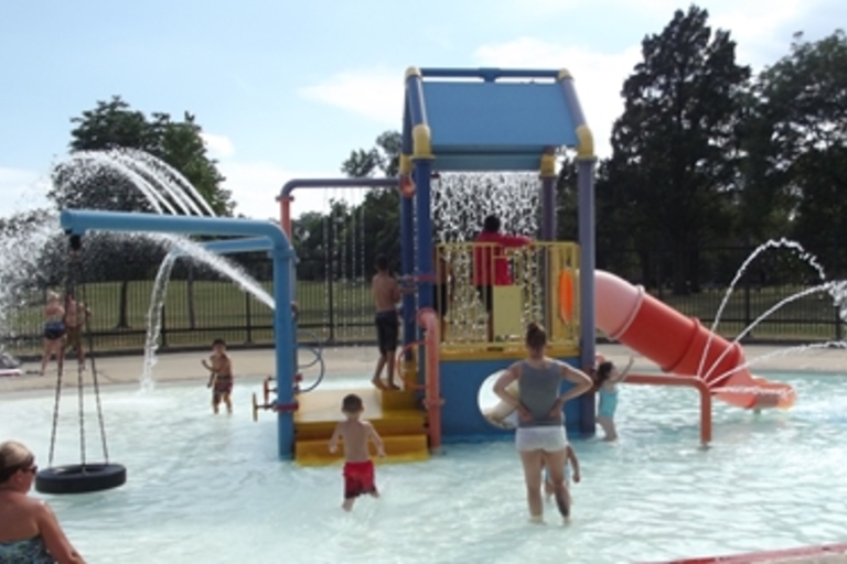 Children and adults play on a colorful splash pad at a water park.