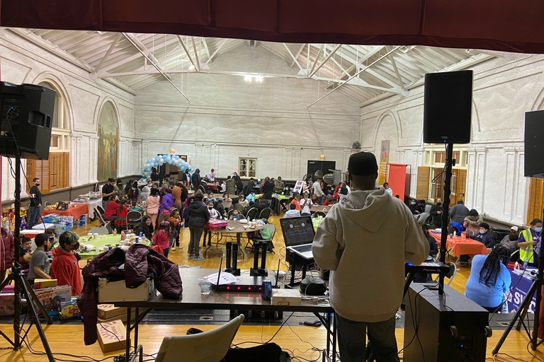 DJ on stage overlooks a large hall with round tables and people.