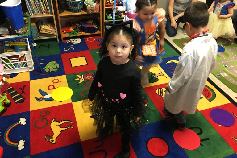 A child in a black cat costume stands on a colorful alphabet rug in a classroom. Other costumed children are slightly blurred in the background.