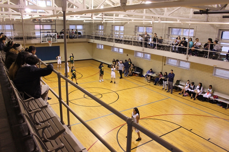 Girls' basketball game in a high school gym. Spectators watch from the balcony.