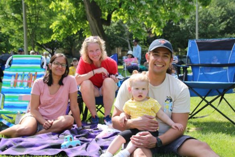 Two women, a man, and a baby sit on a blanket in a park.