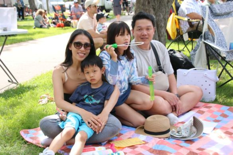 Family of four sits on a blanket at a park.  The daughter blows bubbles.