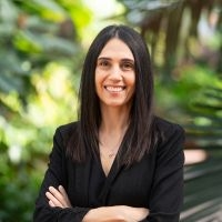 Smiling woman with arms crossed, posing in front of greenery.