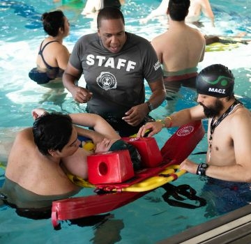 A staff member advises lifeguards as they practice water rescue techniques with a person on a backboard.