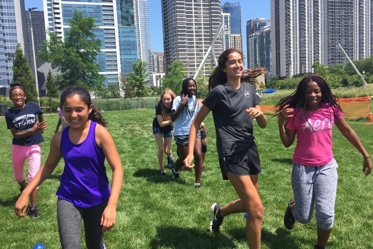 Girls running on grass field with city skyline in background.