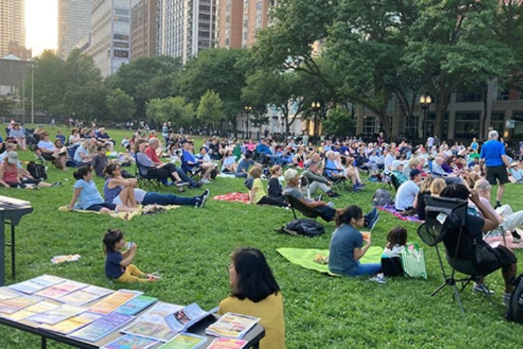 crowd gathered in park in lawn chairs and on blankets, awaiting a performance, city buildings in background