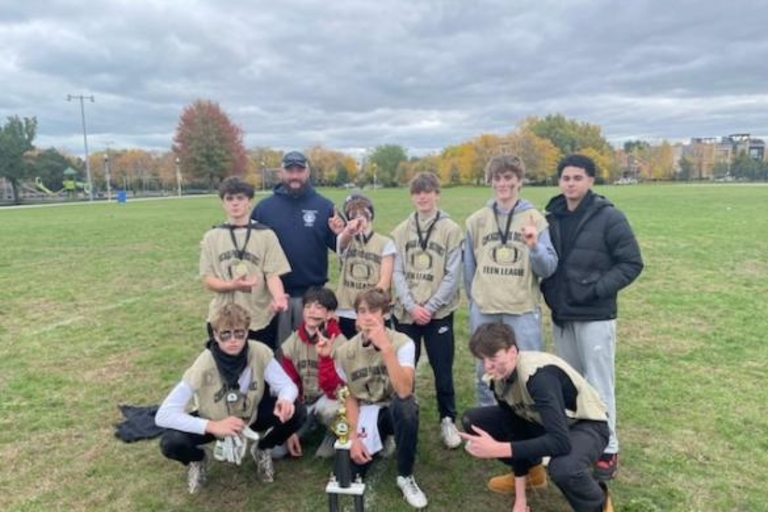Football team and coach pose with trophy and medals in a park.
