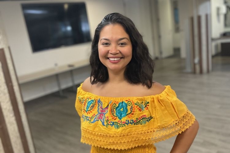 Woman smiling, wearing an off-the-shoulder, embroidered, yellow top.