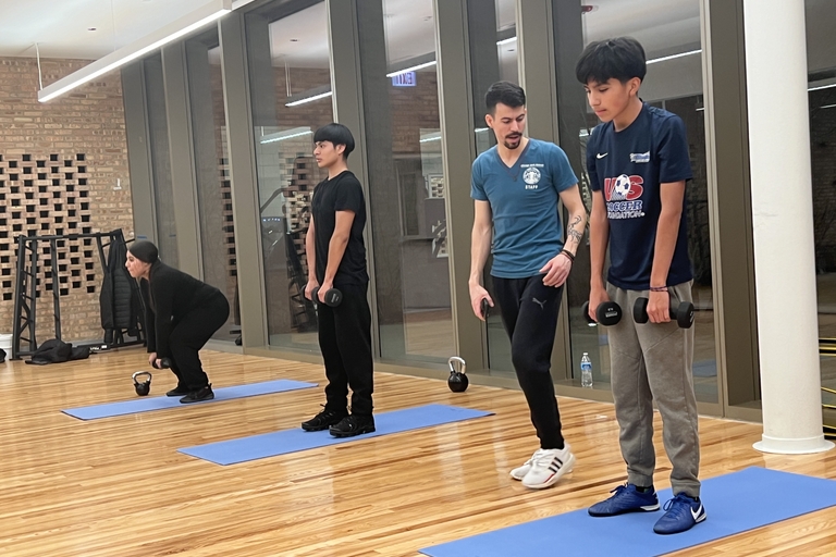 Four people perform dumbbell exercises on blue mats in a gym.