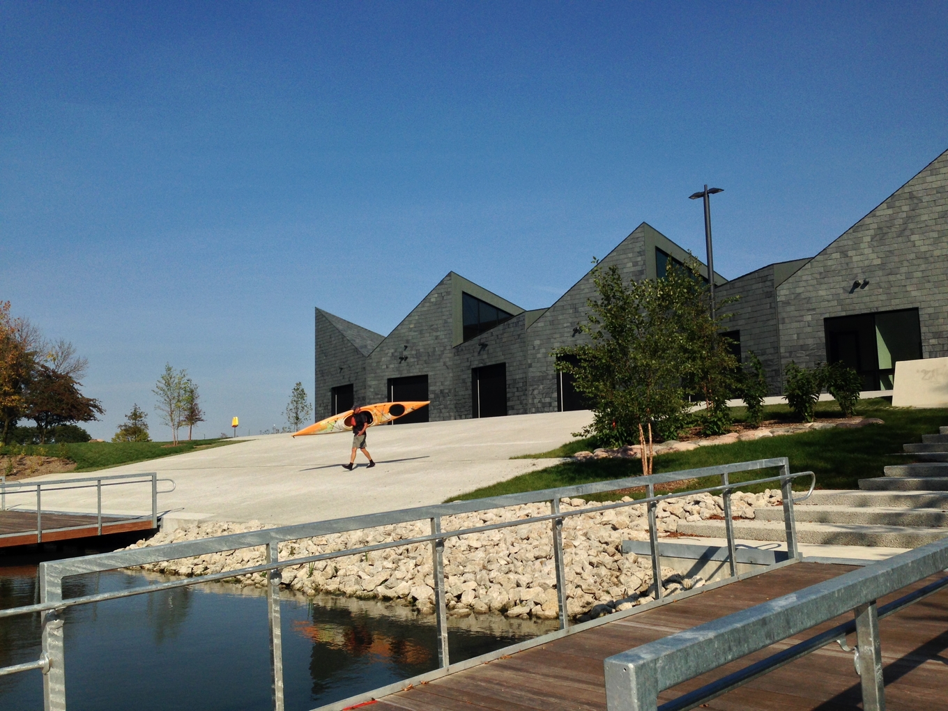 Person carrying kayak towards boathouse on sunny day.