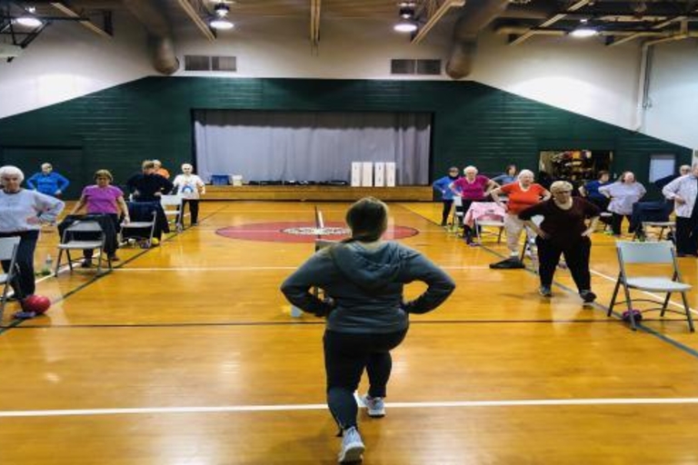 Seniors doing chair exercises in a gym. An instructor faces away from the camera, demonstrating a lunge.