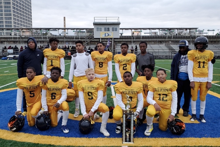 Youth football team in yellow and black Wolverines jerseys pose with a trophy on a field.
