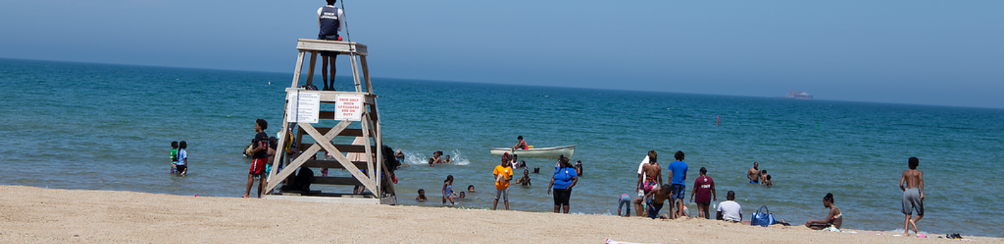 Lifeguard watches swimmers from a chair on a sandy beach.