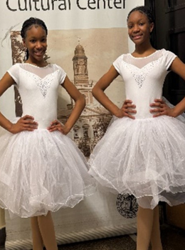 Two smiling ballerinas in white tutus pose before a cultural center backdrop.
