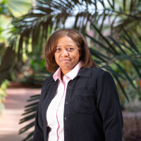 Woman in a black jacket and pink blouse stands in front of palm fronds.