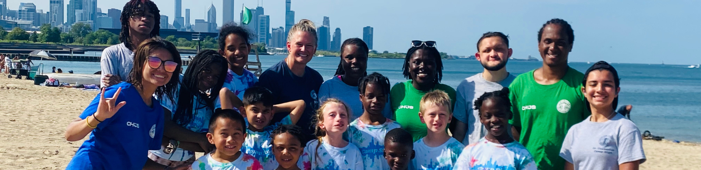 Group photo of children and adults on a beach with a cityscape in the background.