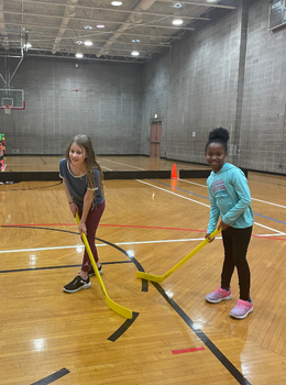 Two girls smile and hold hockey sticks in a gym.
