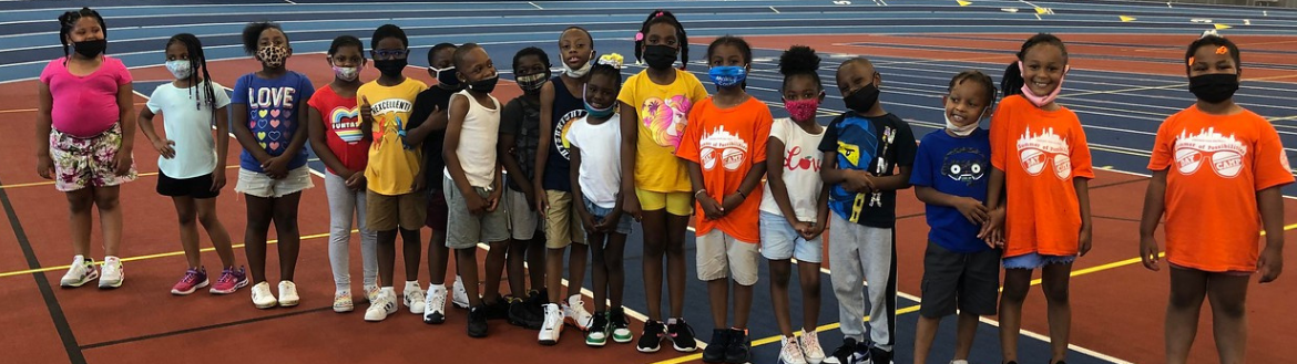 Group of children wearing colorful clothes and masks pose on an indoor track.