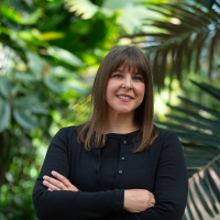 Woman with crossed arms smiles in front of lush foliage.