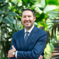 Smiling man in blue suit with arms crossed, posing in front of greenery.